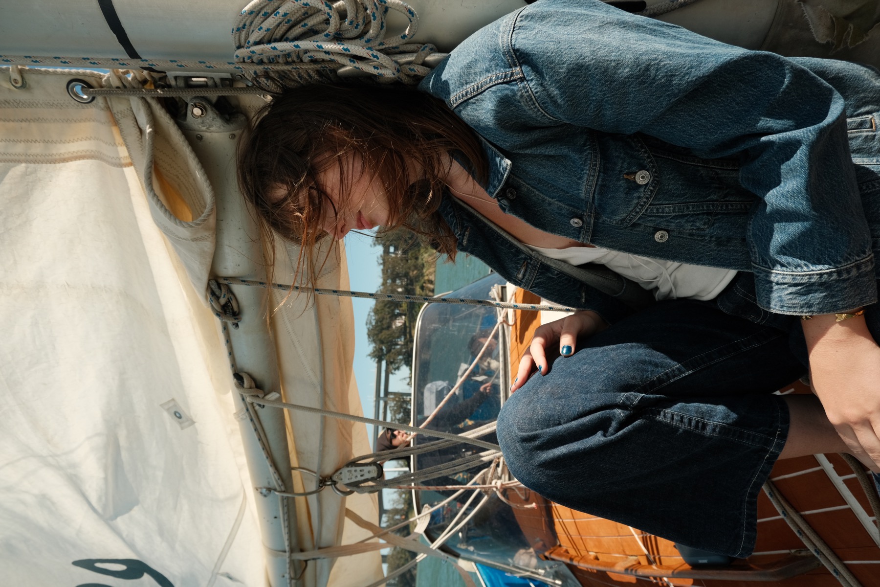 Wind catches a model’s hair beneath a sail, wearing denim on a wooden boat.