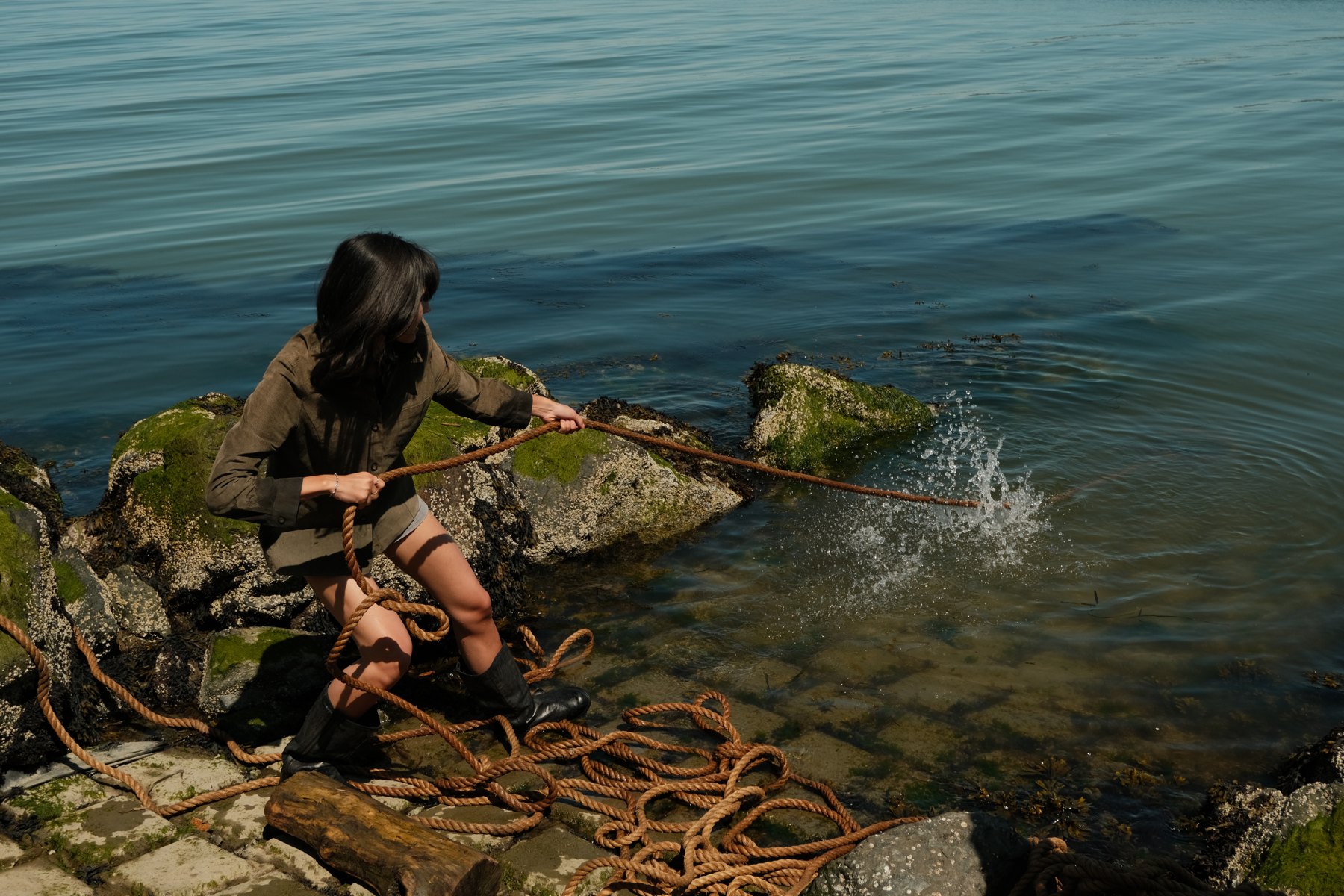 A model among rocks and coiled ropes beside blue water.