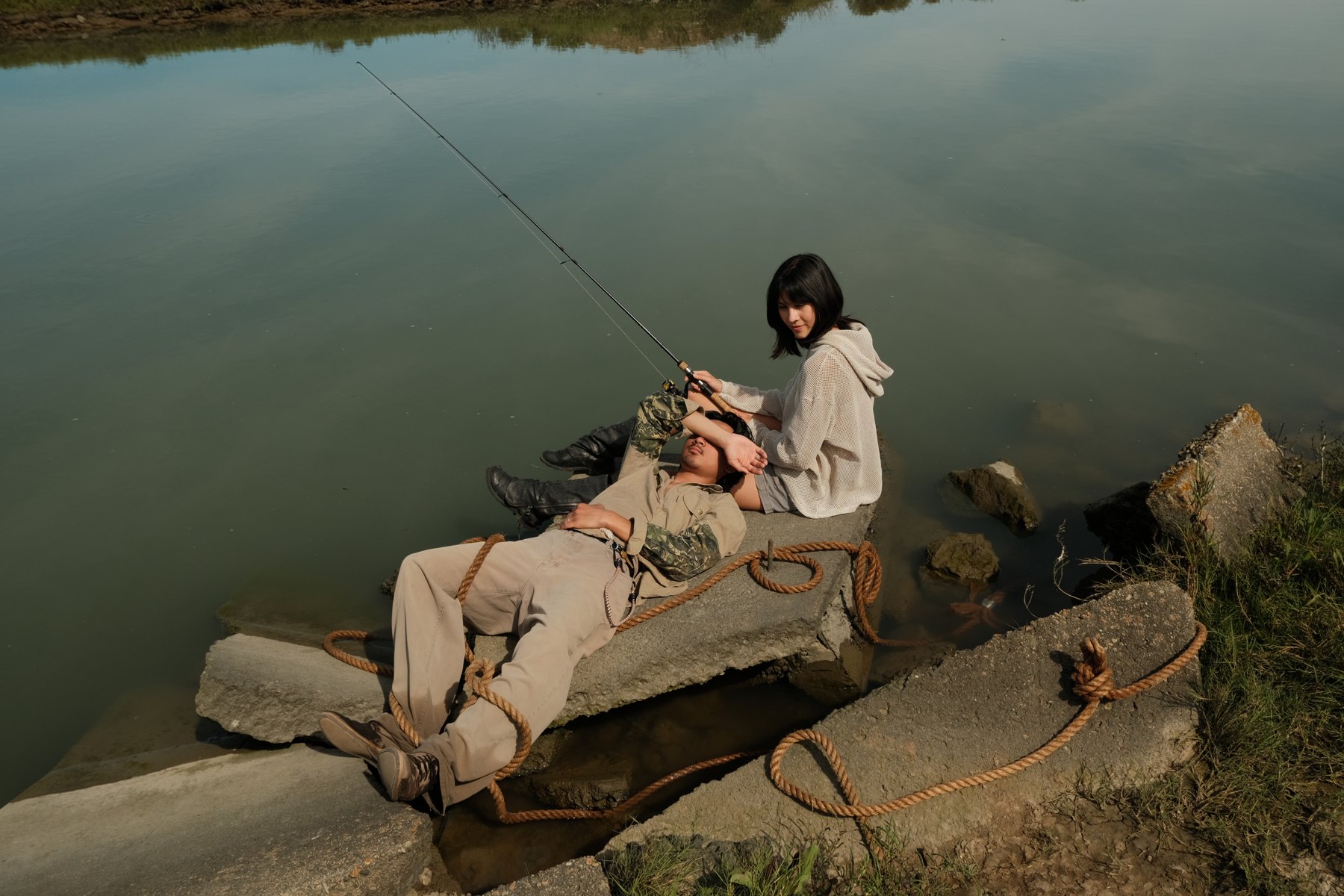 A model seated on rocks by calm water, with rope spread across the stone.