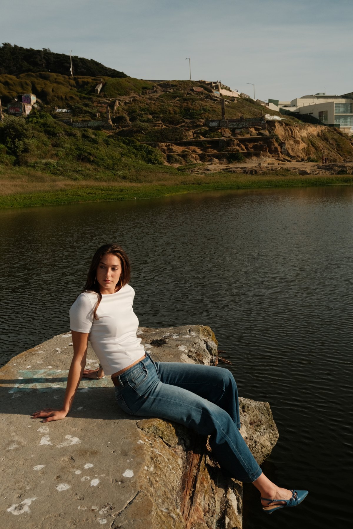 A model wearing denim seated on a stone ledge beside a coastal pool.